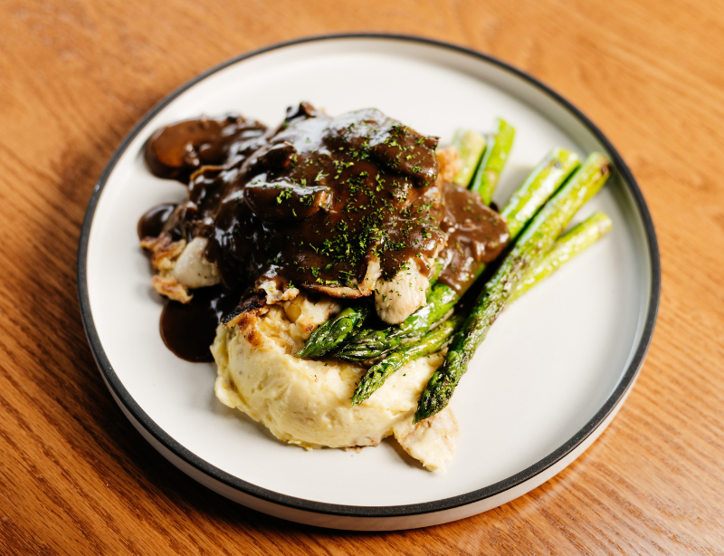 A plate of mashed potatoes topped with grilled chicken, asparagus spears, and dark brown mushroom gravy sits on a wooden table.