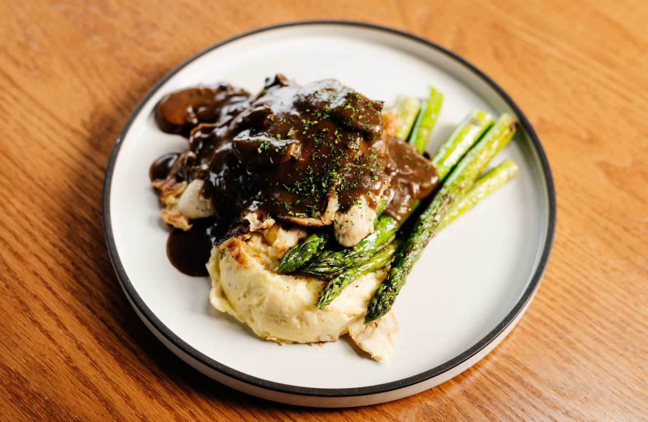 Plate of mashed potatoes topped with chicken, brown mushroom gravy, and herbs, served with roasted asparagus on a white plate with a black rim, placed on a wooden table.