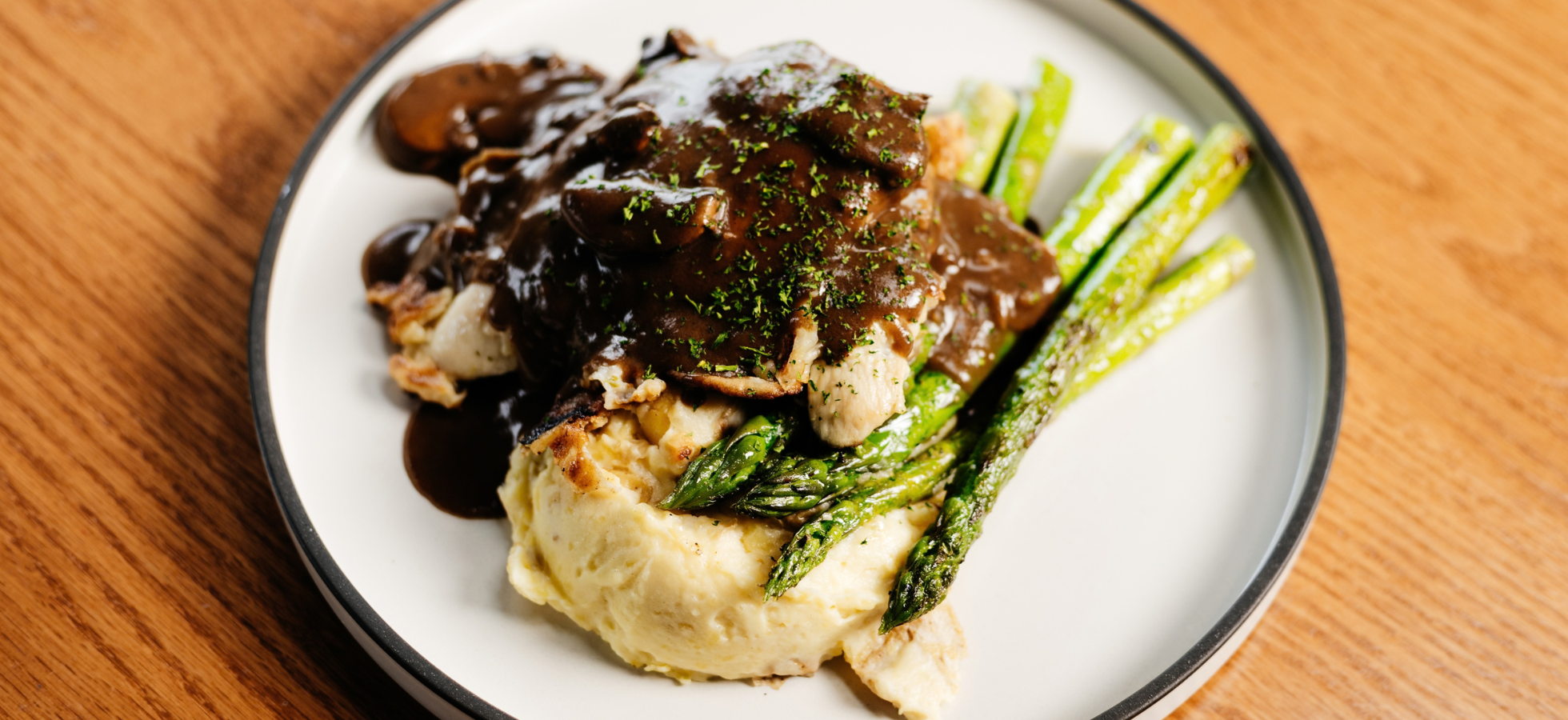 A dinner plate on a wooden table features a serving of creamy mashed potatoes topped with chicken and a dark mushroom gravy, garnished with herbs and accompanied by a side of roasted green asparagus.