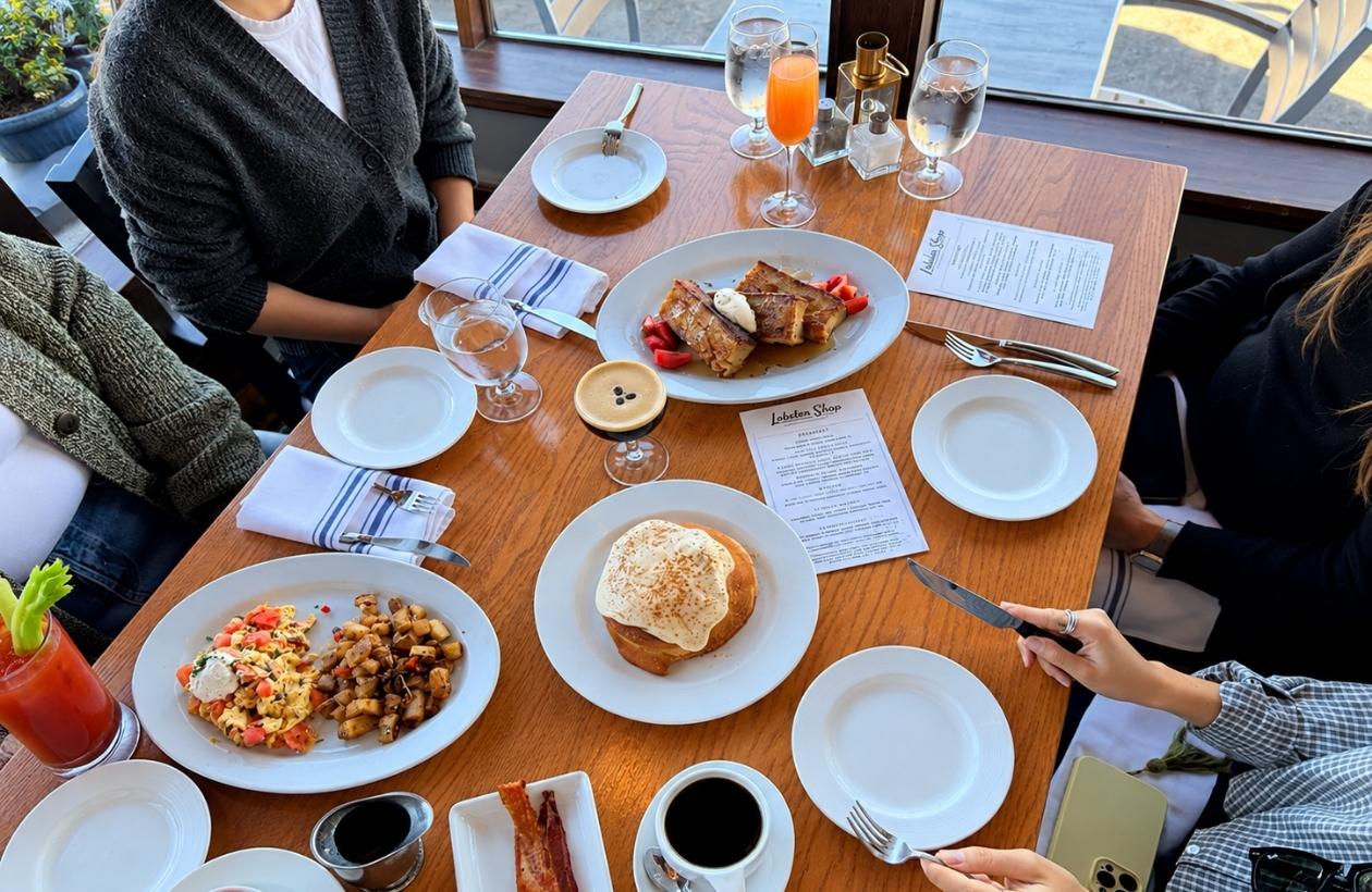 A wooden restaurant table set for four people, featuring plates of breakfast food, cocktails, coffee, and Lobster Shop menus.