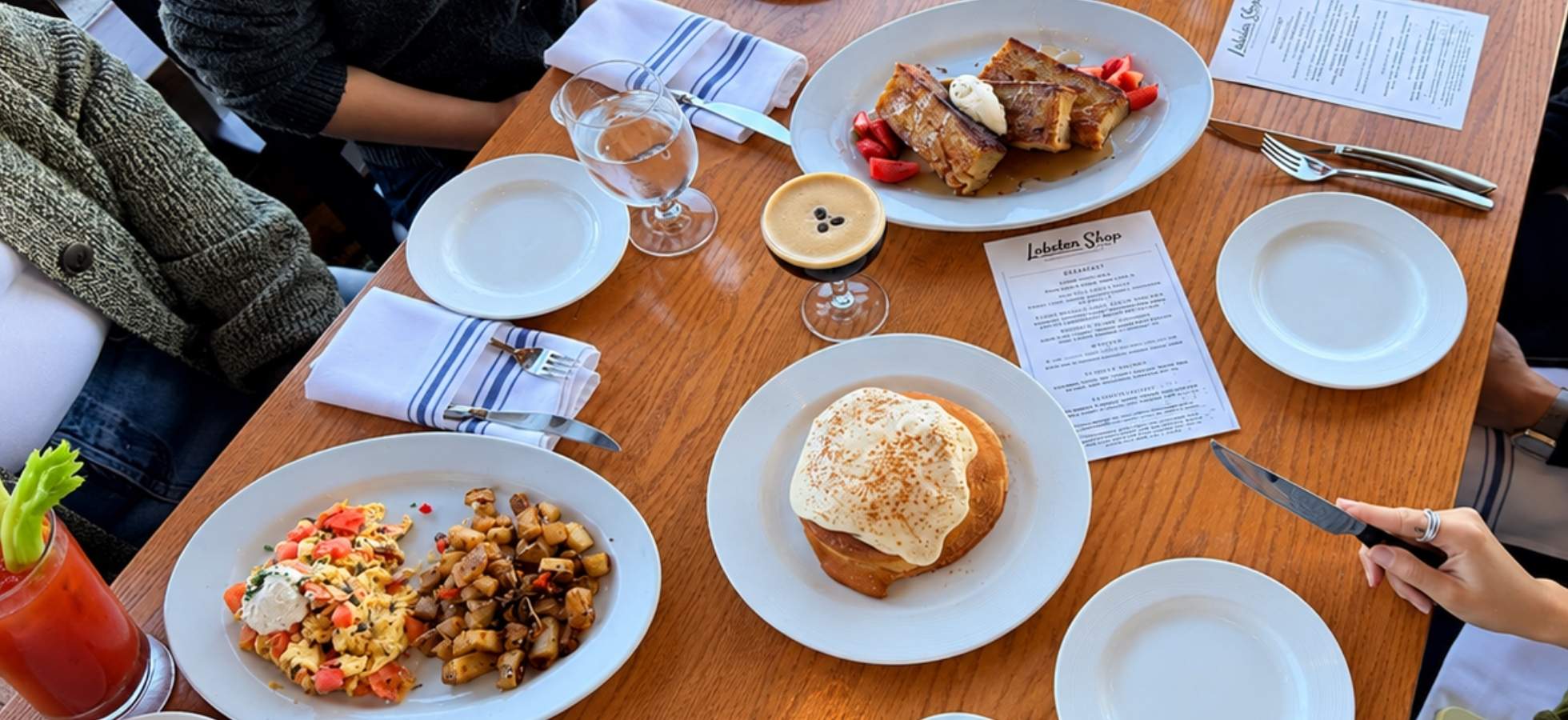 A wooden restaurant table set with plates of scrambled eggs and potatoes, French toast with strawberries, a pastry, an espresso martini, and printed menus.